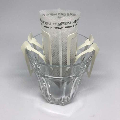 A disposable Beanz Drip bag hanging on a glass cup, ready for hot water to be poured for brewing.