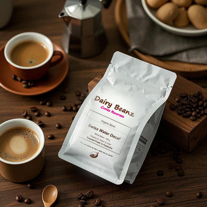 Dairy Beanz Swiss Water Decaf organic espresso blend coffee bag beside two white cups of black coffee with rich crema, a moka pot, scattered beans, and baked treats on a wooden table.