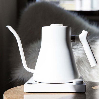A white Fellow Stagg EKG Electric Kettle on the coffee table front the couch pillow at home with sun light.