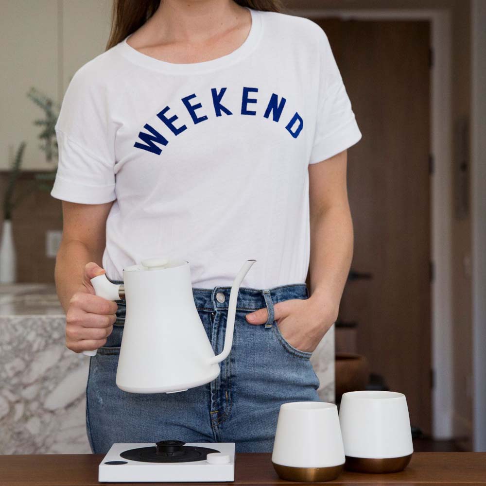 A woman holding a white Fellow Stagg EKG Electric Kettle in front of a table at home, is going to pouring the hot water into two white Fellow cups.