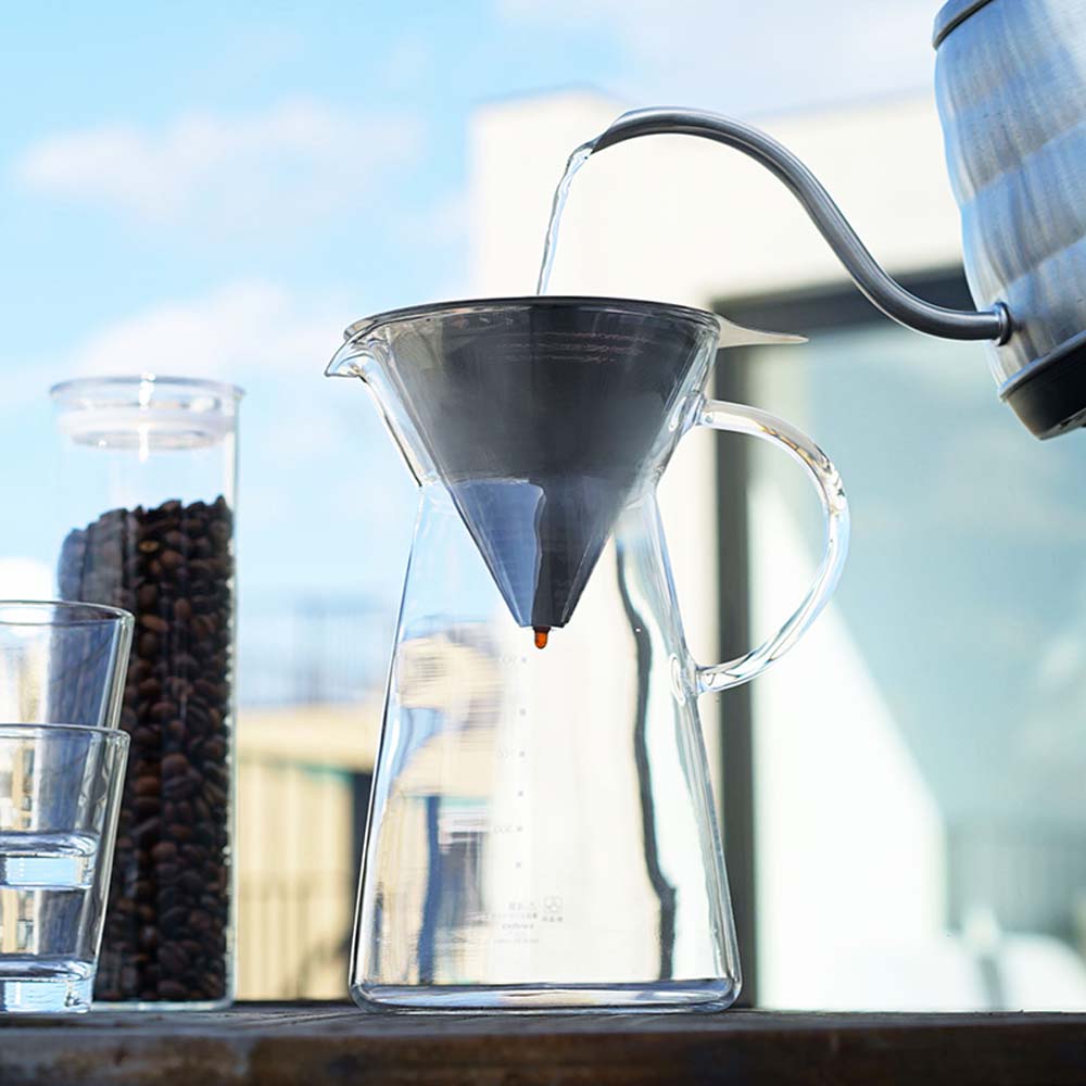 The Hario Metal Dripper with Decanter is an all stainless steel cone dripper that sits over a high-quality borosilicate glass decanter. Pouring hot water and byside two glass cups and a glass tube of coffee beans.