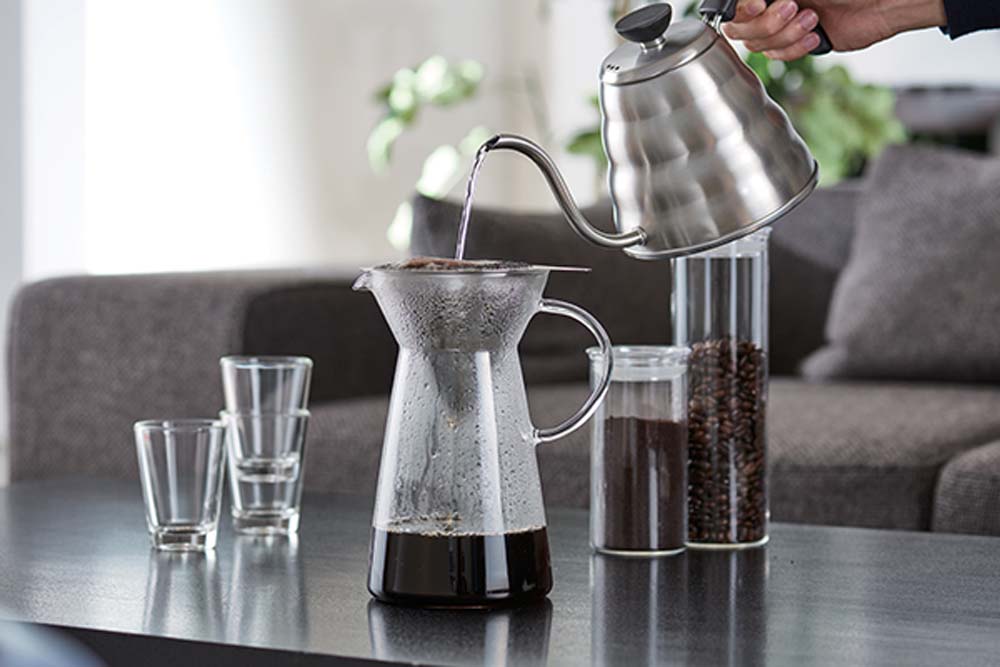 Make coffee at home with the Hario Metal Dripper with Decanter,  Pouring hot water and by side three glass cups and a glass tube of coffee beans and ground coffee.